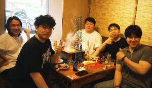 This is a casual group photo of five male colleagues from the operations team enjoying a grilled fish lunch together at a restaurant. They are gathered around the table, smiling brightly at the camera, with several team members flashing thumbs-up gestures. In the center of the table is a steaming grilled fish dish (or hotpot), surrounded by drinks, side dishes, and tableware, creating a warm and appetizing dining atmosphere. To the right, there is a bamboo-woven partition wall, and behind them is a glass window that lets in bright natural daylight. The overall vibe is relaxed, cheerful, and full of team spirit.